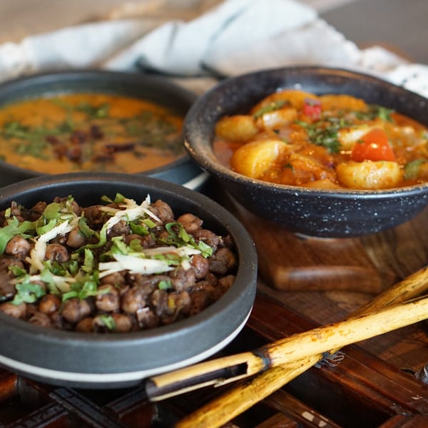 Three bowls of Asian-fusion dishes, including chickpeas, potatoes, and a curry, on a wooden table.