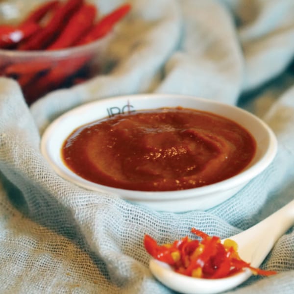 A white bowl of red chili sauce and a spoon with chopped chilies on a light blue cloth.