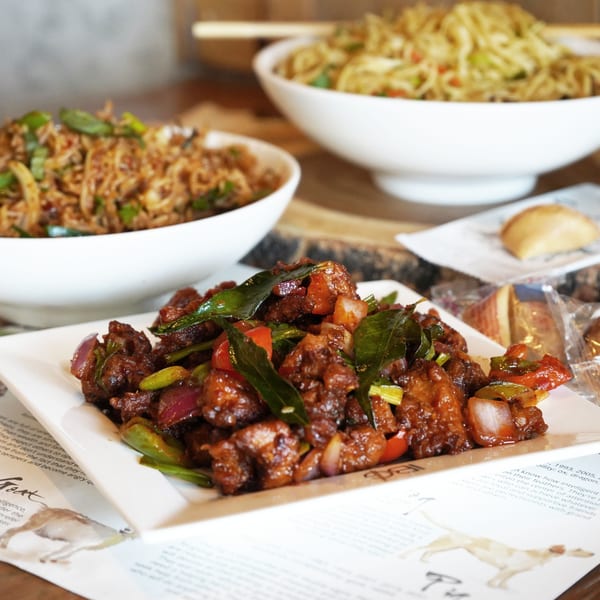 A close-up of chili chicken with bell peppers and onions, with noodles in the background.