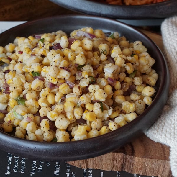 A dark bowl filled with crispy corn kernels, red onions, and green herbs, seasoned with black pepper.