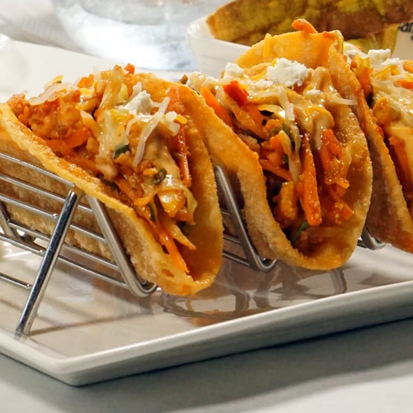 Three Asian-fusion tacos with shredded vegetables, sauce, and cheese, in a metal holder.