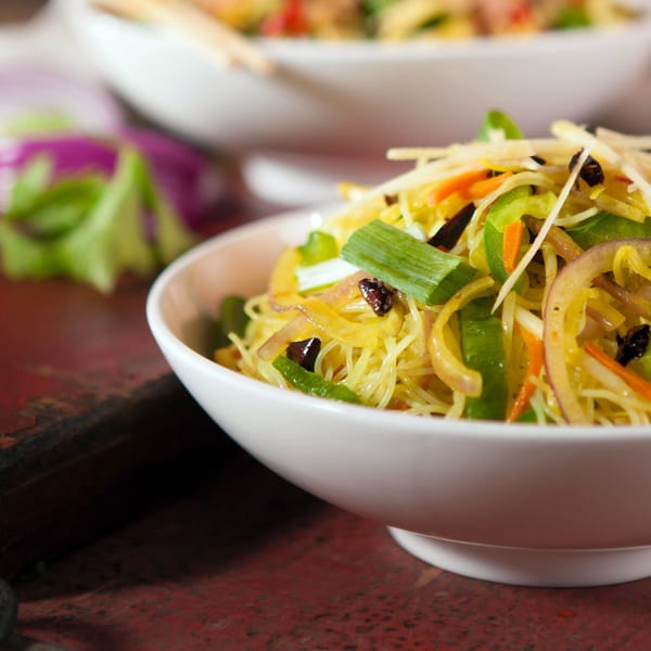Close-up of a white bowl filled with Singapore noodles, vegetables, and dark olives on a rustic red table.