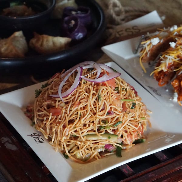 Crispy noodle salad with red onions, tomatoes, and cucumbers, next to tacos and dumplings.