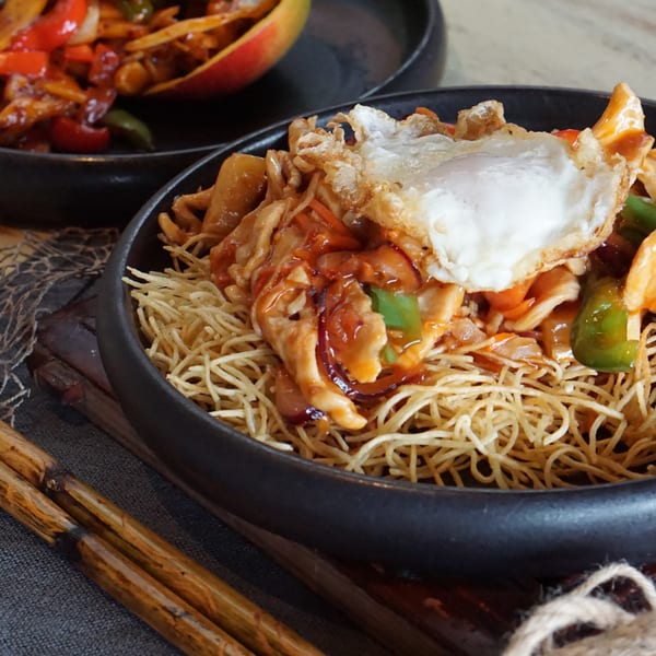 Crispy noodles topped with chicken, vegetables, sweet sauce, and a fried egg, with chopsticks nearby.