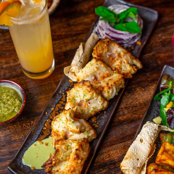Chicken tikka, green chutney, and a cocktail with orange slices on a wooden table.