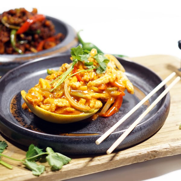 Sweet and sour chicken in a mango half, garnished with cilantro, on a dark plate with chopsticks.